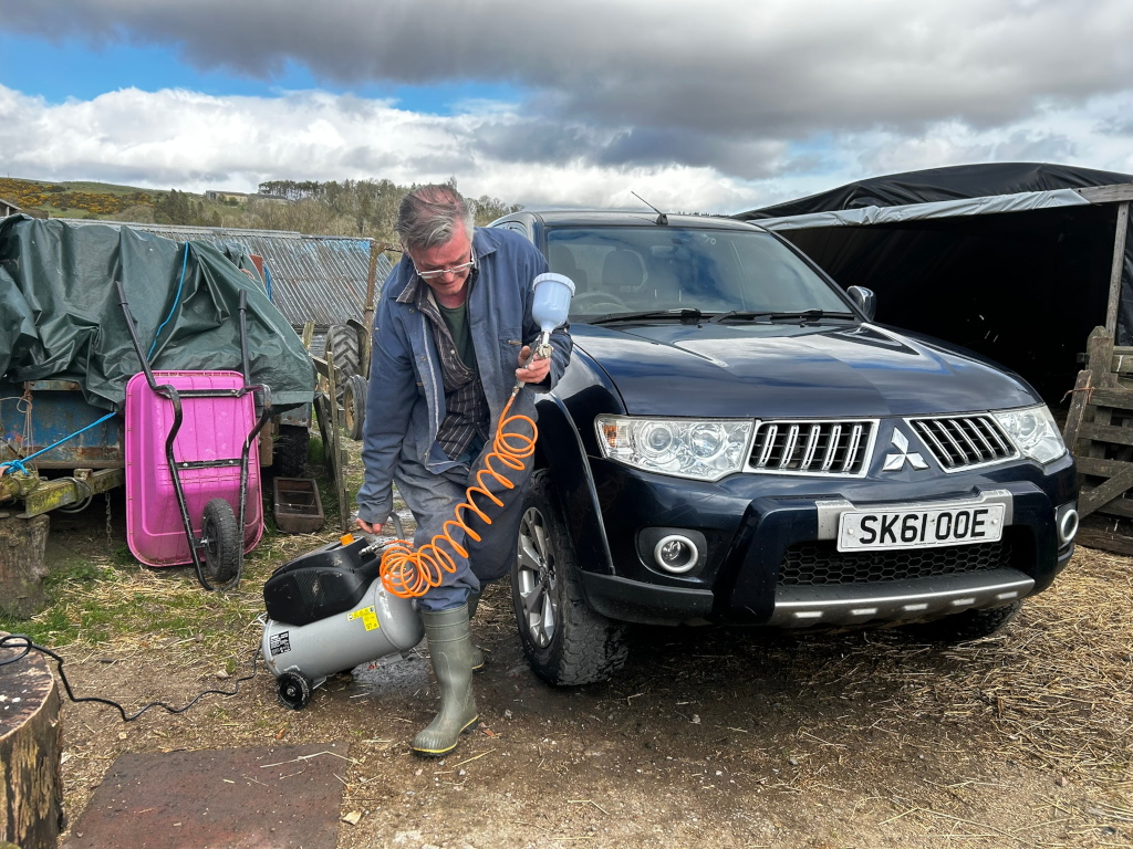 Charlie in work boots and a jacket using an air compressor to spray paint a dark-blue Mitsubishi L200 pickup truck. He's working outdoors on a farm, with a pink wheelbarrow and a tarp-covered trailer visible in the background.