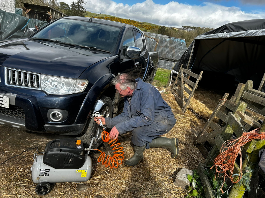 Charlie kneeling beside a dark-coloured pickup truck in a rural setting. He is using an air compressor and spray gun to spray paint to protect the trucks chassis. The background includes farm structures, hay bales, and a partly cloudy sky. The overall impression is one of a farmer performing routine vehicle maintenance in a working farm environment.