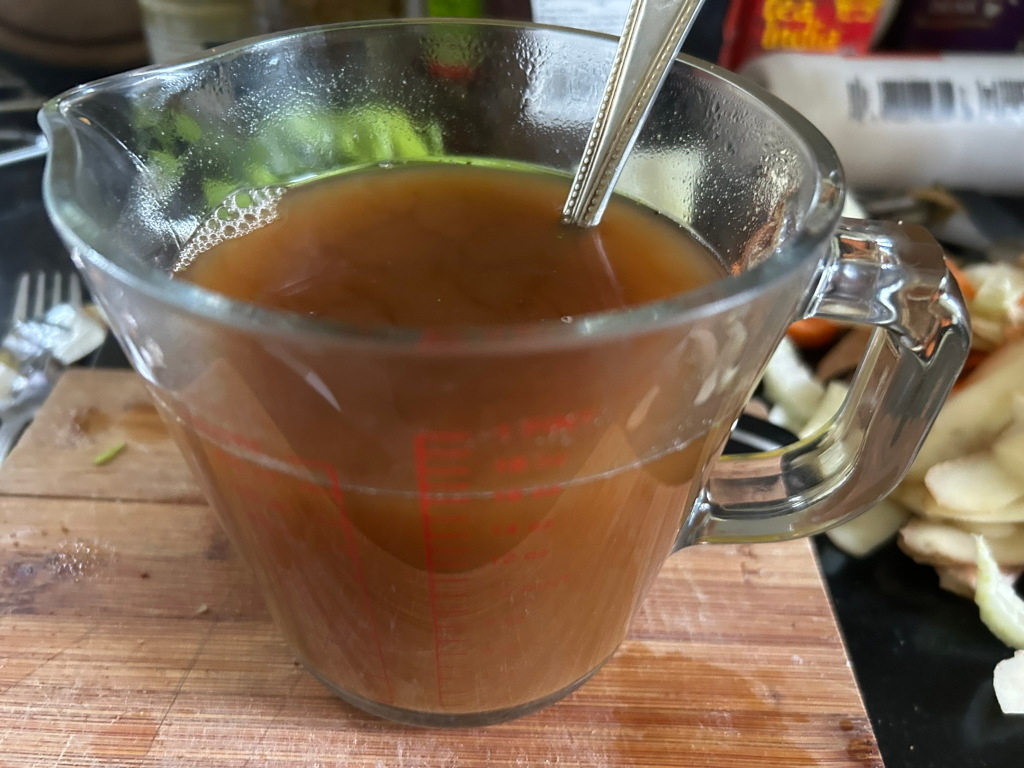 Clear glass measuring cup filled with a dark brown liquid, possibly stock or broth. A spoon is partially submerged in the liquid. A wooden cutting board is visible in the background, along with some chopped vegetables. The overall context suggests a food preparation scene.
