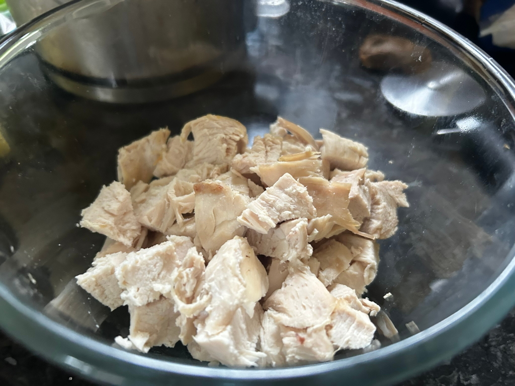 Glass bowl filled with roughly chopped, cooked chicken breast. The chicken appears to be plain, without any visible seasonings or sauces. The lighting is somewhat dim, and the focus is primarily on the chicken itself. There is a slight blurriness to the background suggesting a kitchen setting.