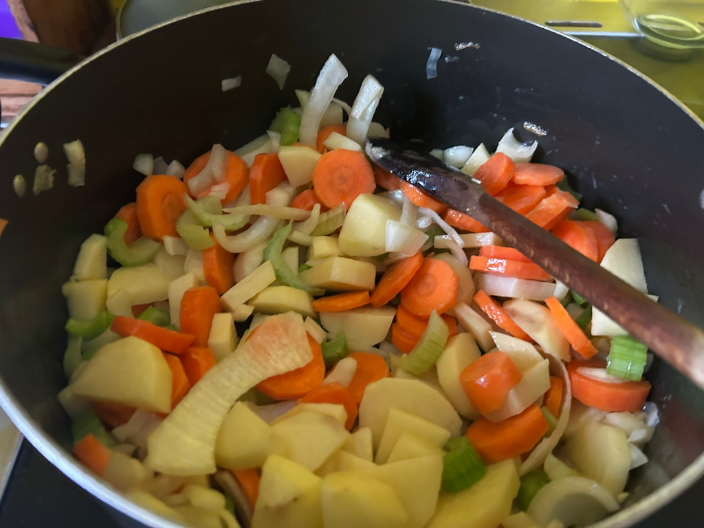 Dark-coloured pot filled with chopped vegetables. The vegetables appear to be carrots, potatoes, onions, and celery, and are being cooked or sautéed. A wooden spoon rests in the pot. The overall impression is of a simple, home-cooked meal in progress.