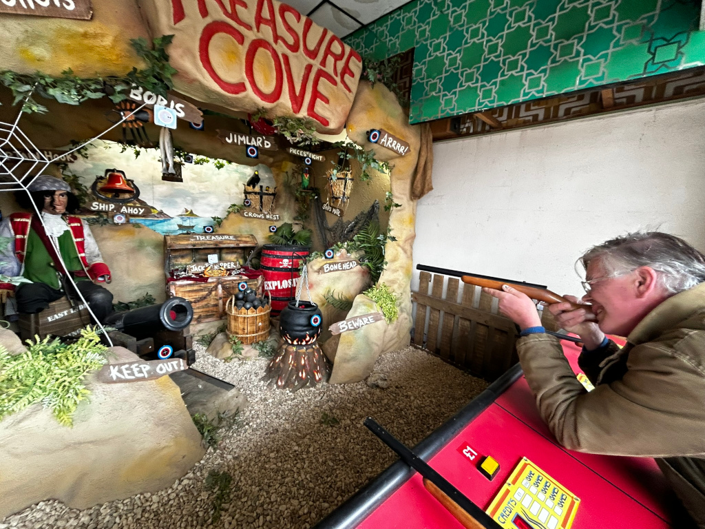 Charlie playing a shooting arcade game themed around a pirate's treasure cove. The game features a detailed diorama with pirate figures, a cannon, treasure chest, and other pirate-themed props. Charlie is aiming a toy rifle at targets within the diorama, seemingly attempting to hit various elements in the scene for points. The creative background is a whimsical, child-friendly depiction of a pirate's treasure hunt. The emotional expression is one of playful engagement; Charlie appears focused and entertained.
