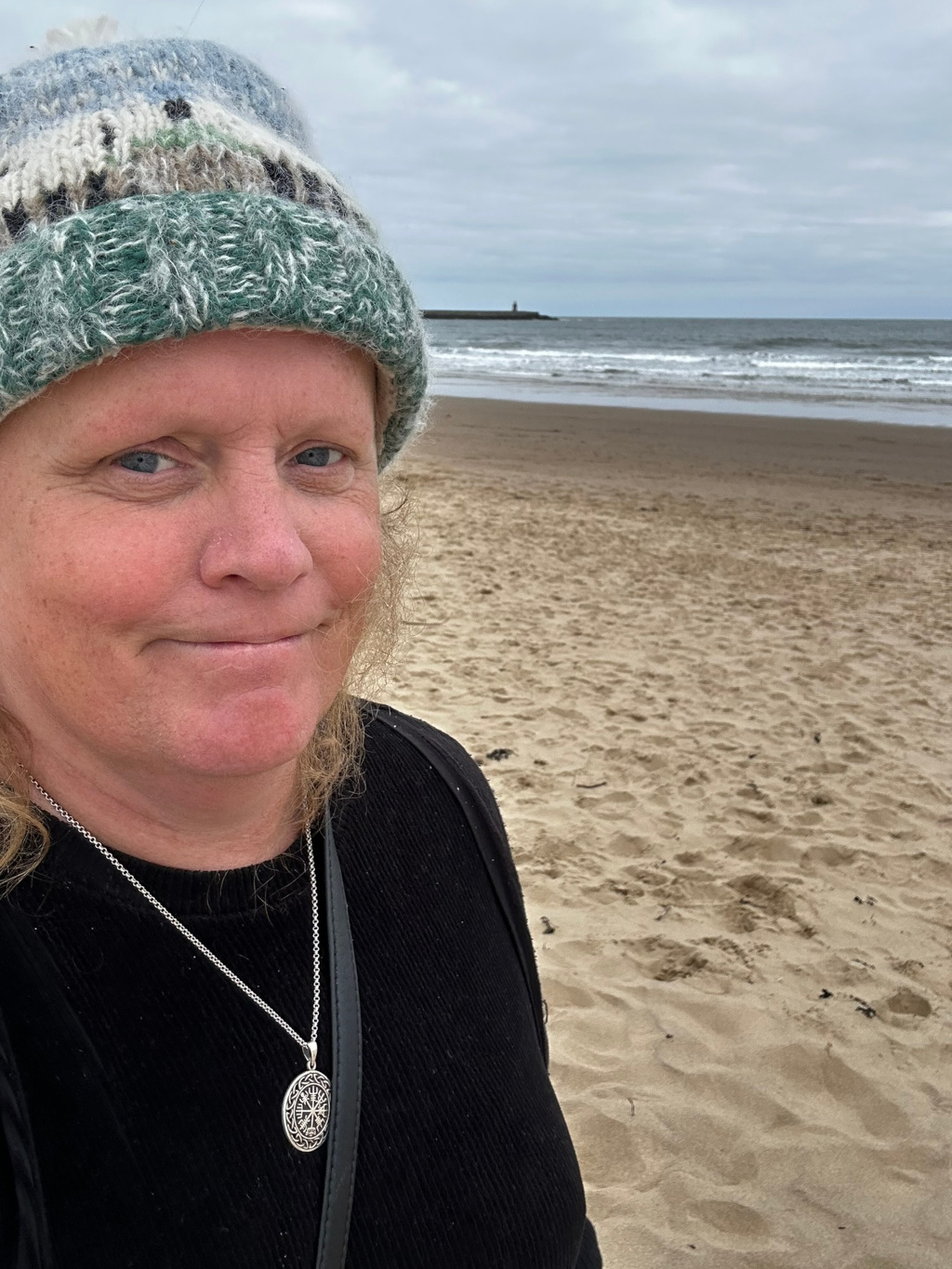 Close-up selfie of a person named Leonie, with fair skin and shoulder-length light brown hair, wearing a hand-knitted green and blue hat and a black sweater. They are wearing a silver necklace with a circular pendant. The background is an out-of-focus sandy beach with a calm sea and a distant pier visible on the horizon under a cloudy sky.