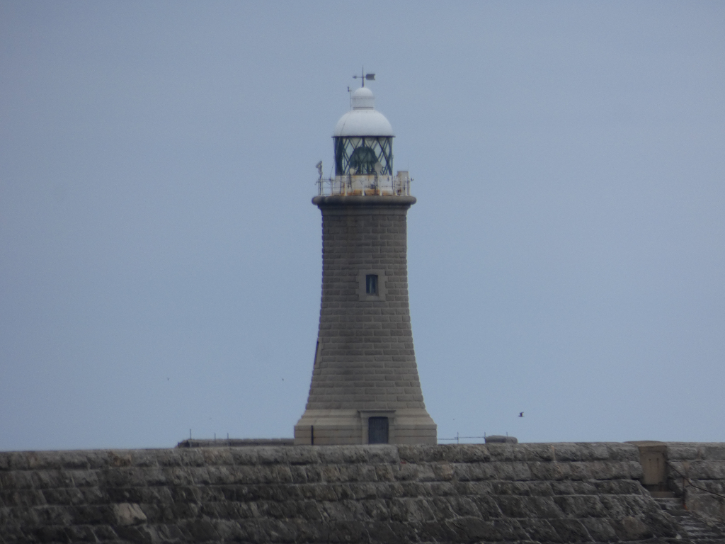 Stone lighthouse situated atop a stone wall, under a muted, overcast sky. The lighthouse is tall and slender, with a white lantern room at its peak. The overall mood is quiet and somewhat austere.