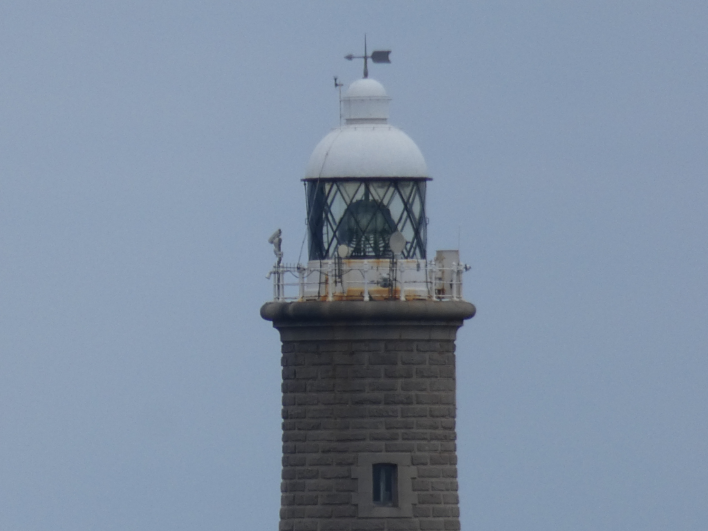 Close-up view of the top of a lighthouse. The lighthouse is made of dark grey stone, and the top section features a white dome covering the lantern room with its glass panes. A weather vane sits atop the dome. There are also some modern technological additions visible near the lantern, likely navigational aids. The sky is a light, uniform blue.