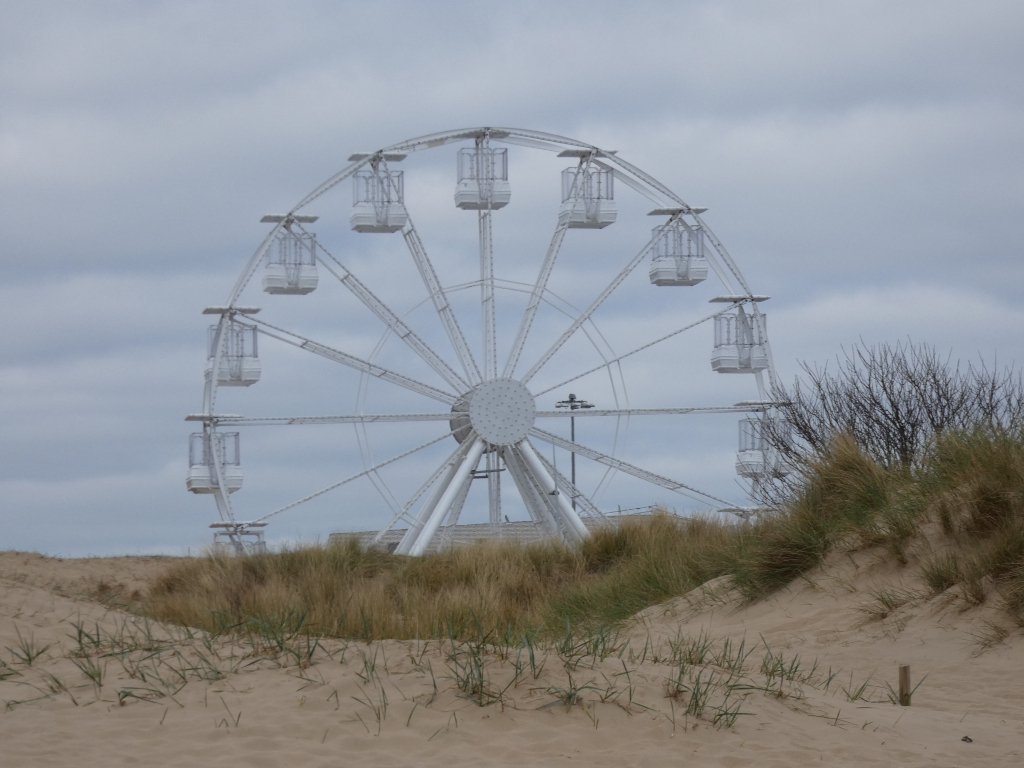 Large white Ferris wheel partially obscured by sand dunes and beach grass. The sky is overcast and gray. The overall impression is one of quiet solitude and a slightly melancholic atmosphere, possibly suggesting an off-season or deserted beach setting. The stark white of the Ferris wheel contrasts with the muted tones of the sand and sky.