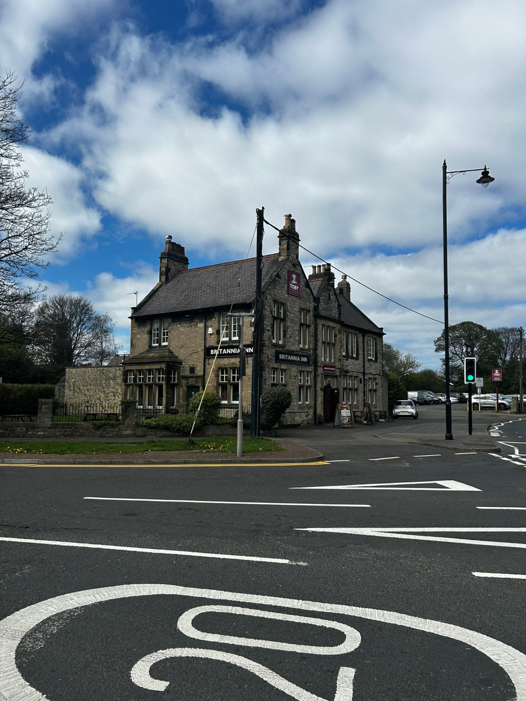 Britannia Inn, a stone pub, situated on a corner where two roads intersect. The pub is a two-story building with traditional architecture.  The sky is partly cloudy, with a mix of blue sky and fluffy white clouds. There are cars parked nearby, and a traffic light is visible at the intersection. The overall scene suggests a quiet, possibly suburban, location in the UK.