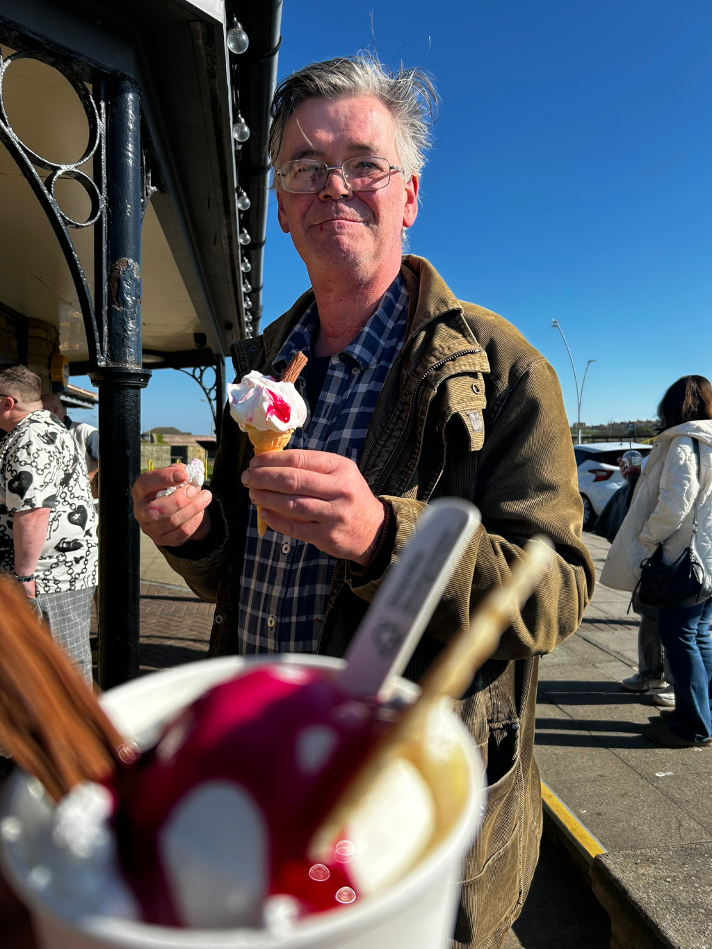Charlie with glasses and slightly windblown gray hair, smiling at the camera while holding an ice cream cone in his hands. He is wearing a brown corduroy jacket over a plaid shirt.  In the foreground, out of focus, is another ice cream dessert in a cup. The background features a seaside promenade setting, with other people visible in the distance, suggesting a sunny and pleasant day. The overall impression is one of casual enjoyment and relaxed happiness.