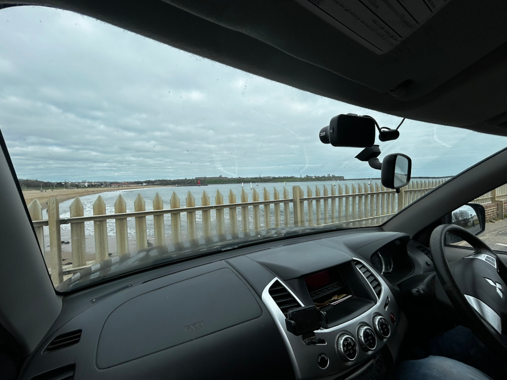 View from inside Charlie's pick up truck, parked near a beach. A white picket fence separates the car from the sandy beach and the calm sea beyond. In the distance, there's a coastline with buildings and a few sailboats visible on the water. The sky is overcast with a grey, cloudy sky. A dash-cam is visible mounted on the car's wind-shield. The overall feel is one of a somewhat gloomy, but peaceful coastal scene.