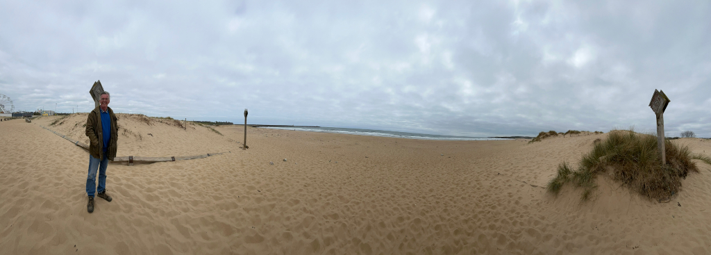 Panoramic view of a sandy beach on an overcast day. Charlie is standing in the foreground near a wooden post and what appears to be a partially damaged boardwalk. In the distance, there's a calm sea and a hint of a Ferris wheel suggesting a nearby amusement park. The overall mood is serene yet somewhat melancholic due to the cloudy sky. The sand dunes flanking the beach add a natural, textured element to the scene. The photo seems to capture a moment of quiet observation or reflection by the man at the edge of the beach.