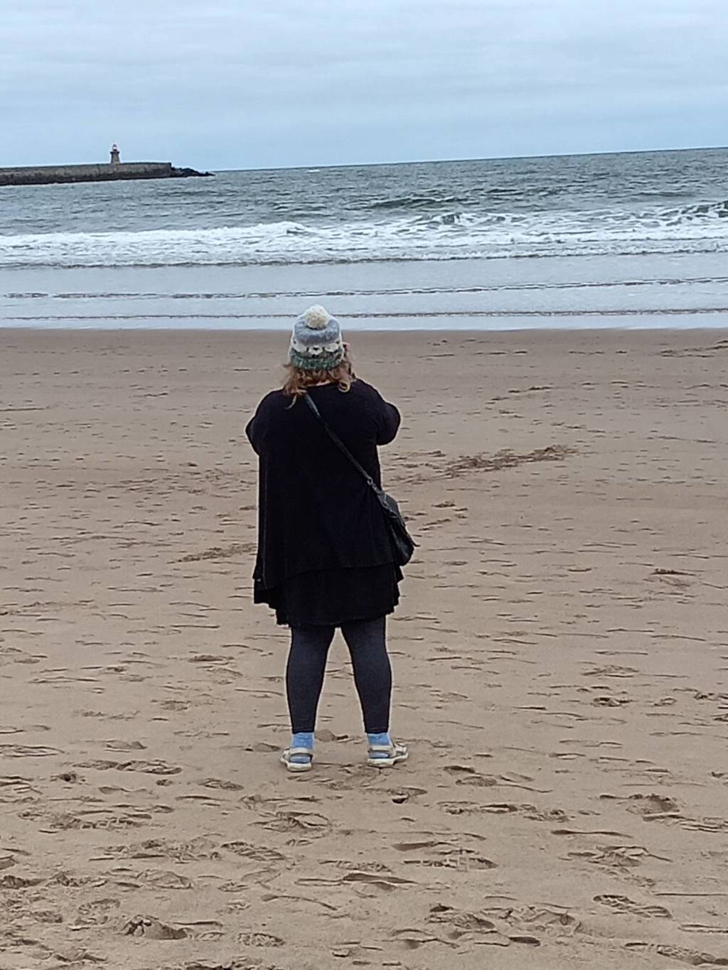 Leonie seen from behind, standing on a sandy beach and facing the sea. She is wearing a dark coat, leggings, and light Coloured shoes, and a knitted hat. Leonie appears to be taking a photograph of the ocean and a distant pier with a small lighthouse. The ocean is relatively calm with small waves.