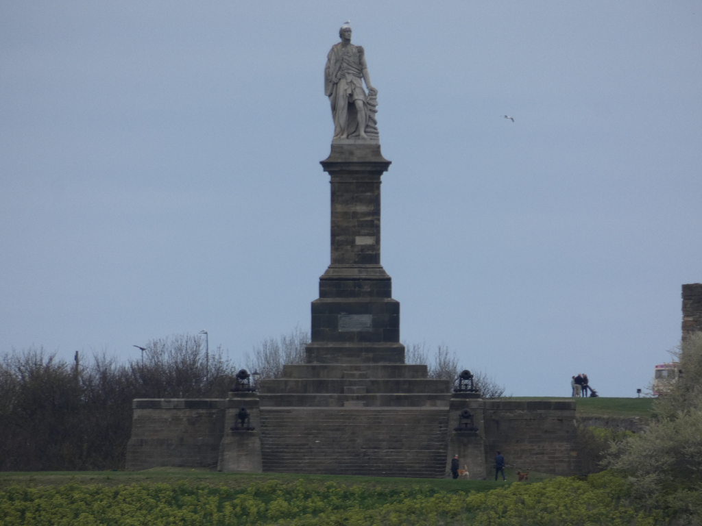 Tall, stone monument featuring a statue of a robed figure atop a multi-tiered pedestal. The monument stands on a raised platform with additional architectural elements at its base. The background includes a muted sky and some sparse trees, indicating a possibly park-like or historical setting. A few small figures of people and dogs can be seen in the distance, giving a sense of scale. The overall aesthetic is sombre and classical in style.