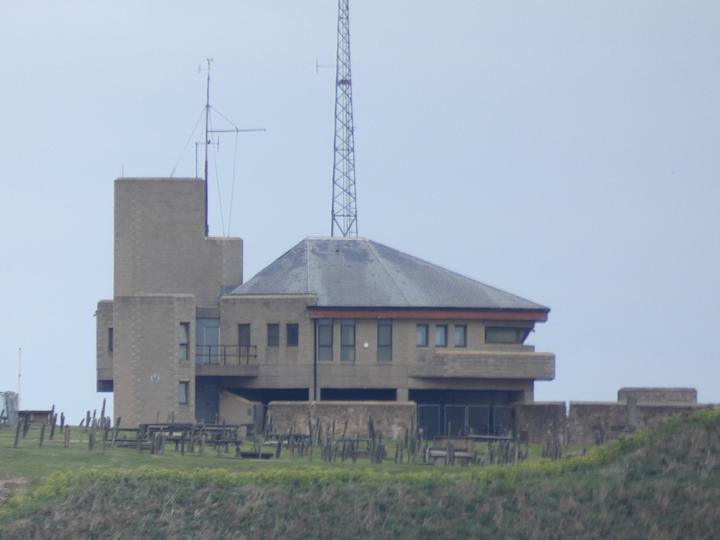 Tan-Coloured, modern building with a sloped roof situated on a hill. The building appears to be a multi-level structure with several windows and a small balcony or terrace. There are various antennas and radio equipment visible on the roof. In the foreground, there's a grassy hill with a few picnic tables.
