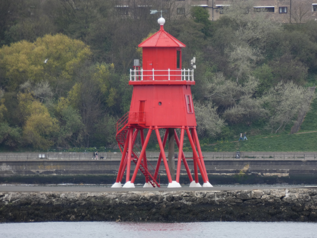 Bright red lighthouse standing on stilts in a body of water.  It's a relatively small structure with a white railing and a spiral staircase on the outside. Behind the lighthouse is a low, rocky seawall and a background of trees and a distant building. A few people are visible walking along the seawall in the distance.