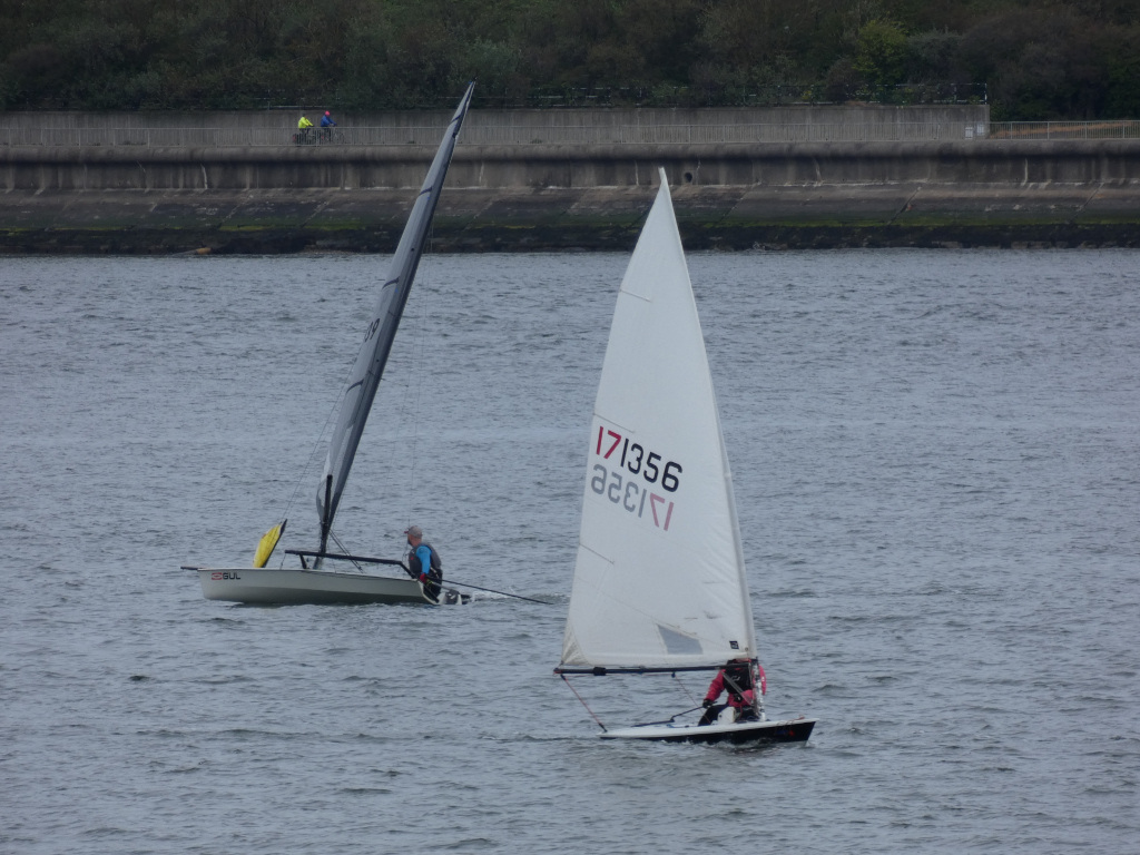 Two sailboats on a body of water. One sailboat is dark-Coloured and has a single person sailing it. The other sailboat is light-Coloured, and also has a single person sailing it. In the background, there is a concrete wall and some trees, and two people are visible walking along the wall.
