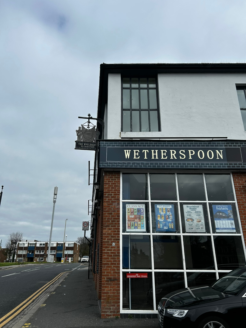 A Wetherspoon pub called The Wouldhave in the UK. The photo is taken from across the street, showcasing the pub's exterior. The building is brick with large windows displaying advertisements for various drinks and breakfast deals offered inside. A prominent Wetherspoon sign is visible, along with a more traditional pub sign above it.