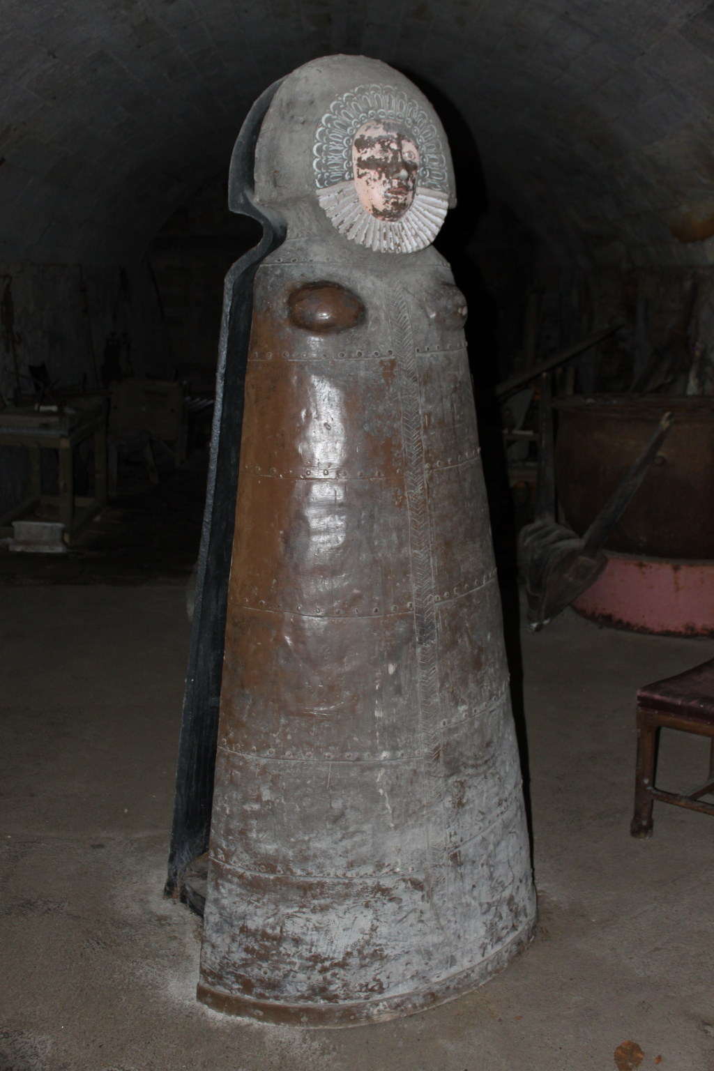 Large, weathered copper sculpture resembling a woman in a long dress or robe. The sculpture's head is a separate element, appearing as a sculpted face with a ruff, affixed to the top of the copper body. The sculpture is situated in what appears to be a dimly lit, possibly underground, space with other indistinct objects in the background suggesting a workshop or storage area.