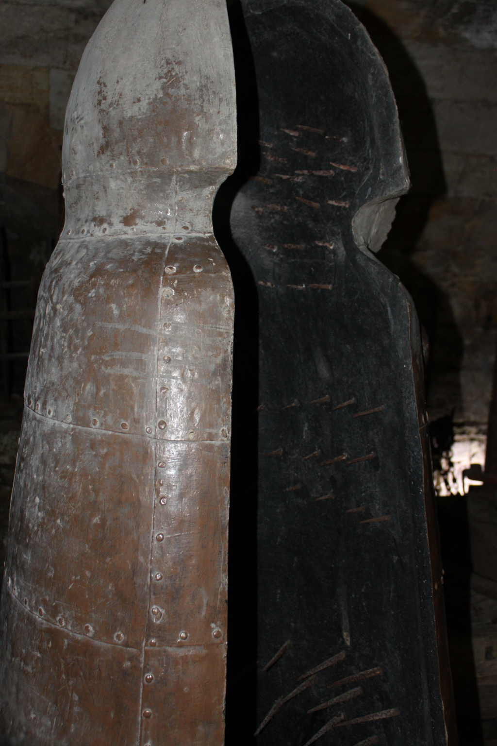 Medieval torture device, specifically a type of iron maiden. The device is depicted open, revealing the interior lined with numerous rusty nails. The exterior is aged and shows signs of wear, further emphasising its historical context and the brutality it represents. The dark, stone setting adds to the grim atmosphere.