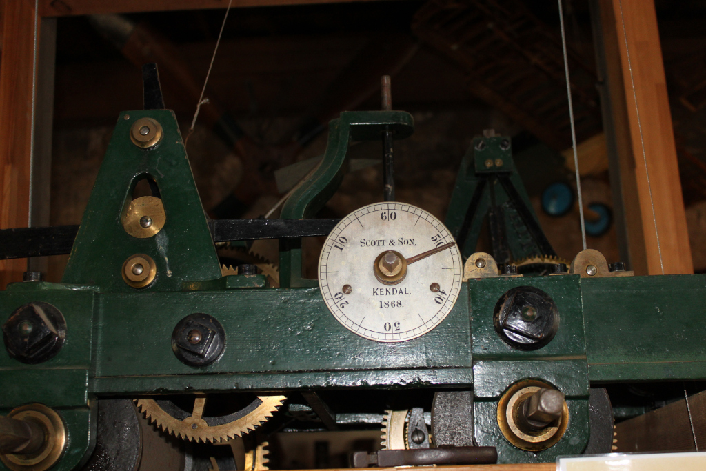 Close-up view of a section of an antique clock mechanism. The mechanism is dark green, with brass gears and other components visible. A prominent circular dial displays the maker's name cott & Son, the location Kendal, and the year 1868. The overall impression is one of age, craftsmanship, and precision engineering.