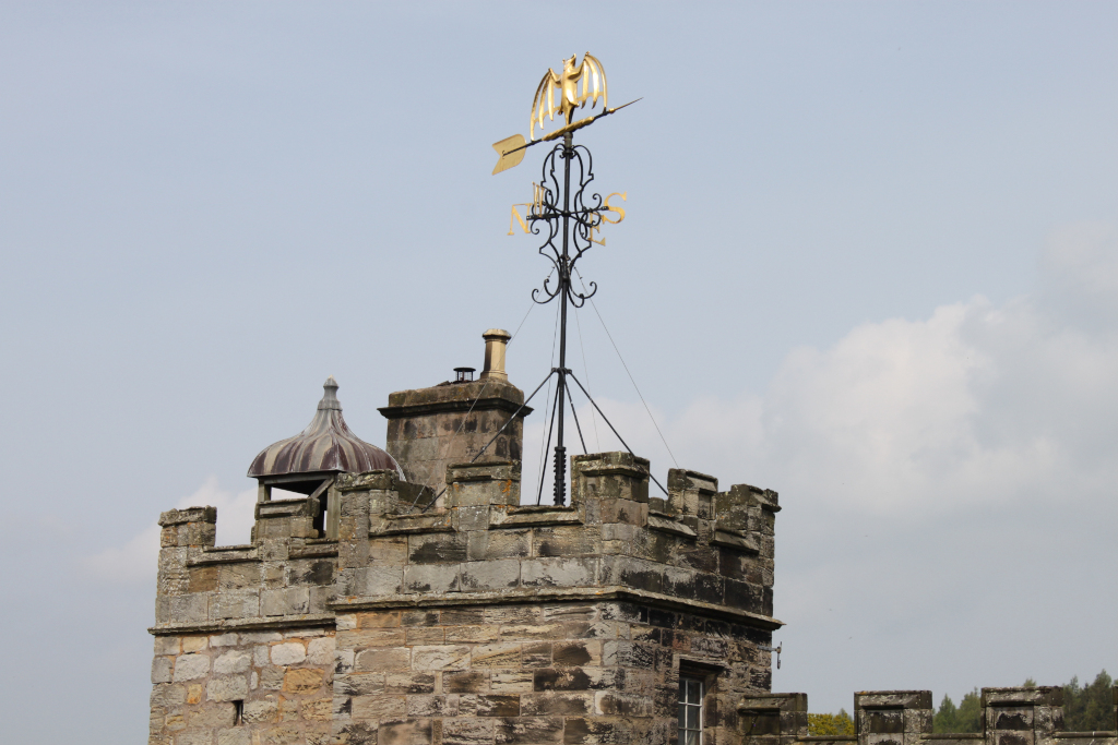 Close-up view of a stone structure, possibly part of a castle or fortified building, topped with a distinctive weather vane. The weather vane is composed of a gold-coloured bat or similar creature design atop a black metal framework, indicating direction. The stonework appears aged and weathered, hinting at historical significance. A smaller, domed structure is visible just below the weather vane, and the overall architectural style suggests a traditional, possibly medieval or early modern, building. The sky is mostly clear with a few clouds.