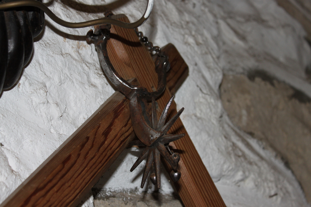 Rusty, antique spur hanging on a wooden cross-shaped structure against a whitewashed stone wall. The spur is the central focus, displaying significant wear and age. The wooden structure appears weathered as well, adding to the overall sense of antiquity. Part of another object, possibly a piece of old machinery or equipment, is visible in the upper left corner.