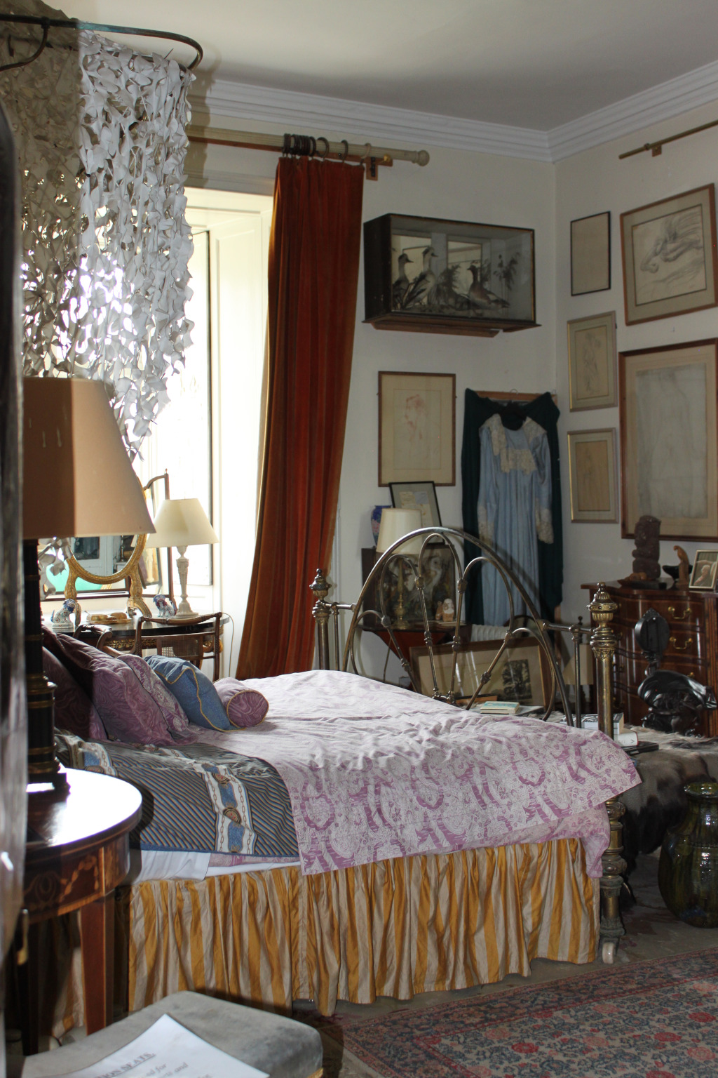 Bedroom with a bed as the focal point. The bed has a metal frame, a patterned comforter, and a gold dust ruffle. Several pieces of artwork and other personal objects decorate the walls and surfaces in the room which appear to be eclectically styled and possibly belonging to an artist.