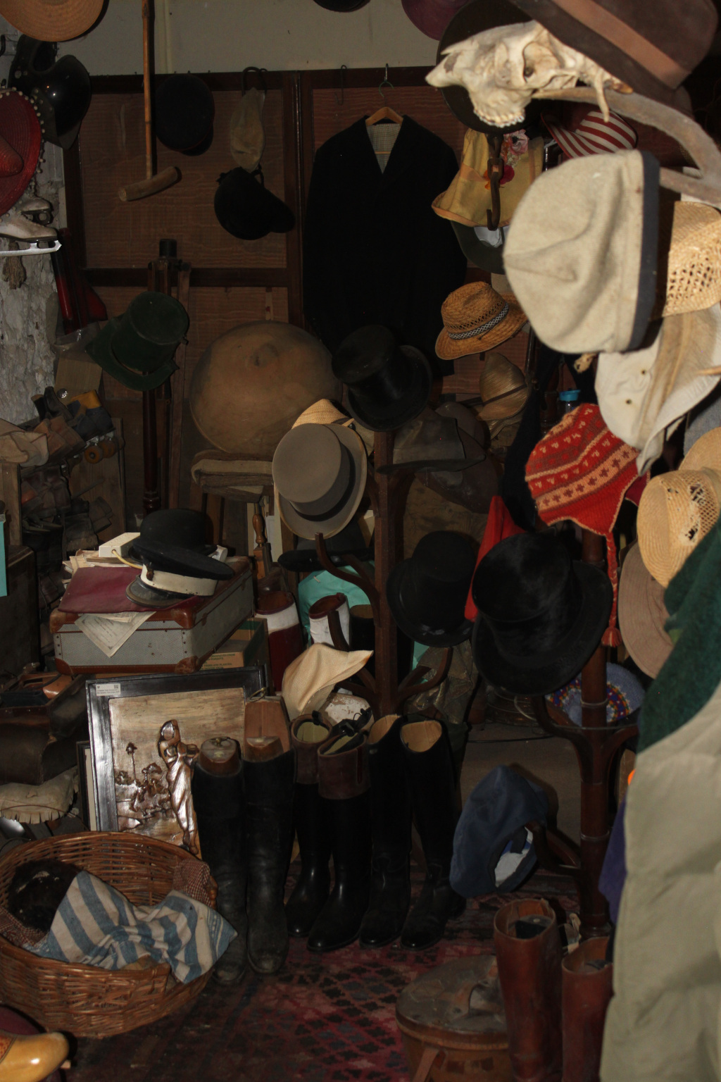 Cluttered room filled with various hats, boots, and other items. Many hats of different styles and materials hang from a rack and are piled on surfaces. Several pairs of riding boots are stacked on the floor. There's a general sense of disorganisation and a feeling of age and history to the objects. The scene suggests a storage room or attic filled with antique or vintage clothing and accessories.