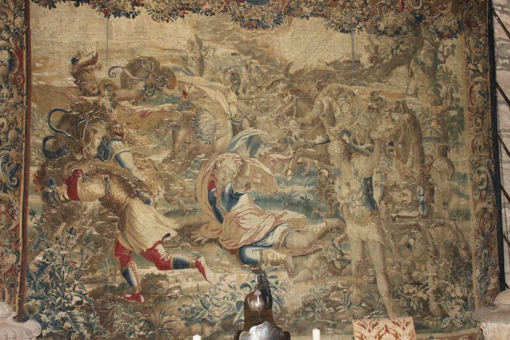 Tapestry depicting a chaotic battle scene. Figures, appearing to be warriors in classical-style attire, are engaged in violent conflict. The style suggests a historical or mythological subject, possibly from antiquity. The tapestry is aged and shows significant wear and tear, adding to its historical feel. The colors are muted earth tones with some faded brighter accents. The overall impression is one of action, violence and the passage of time.
