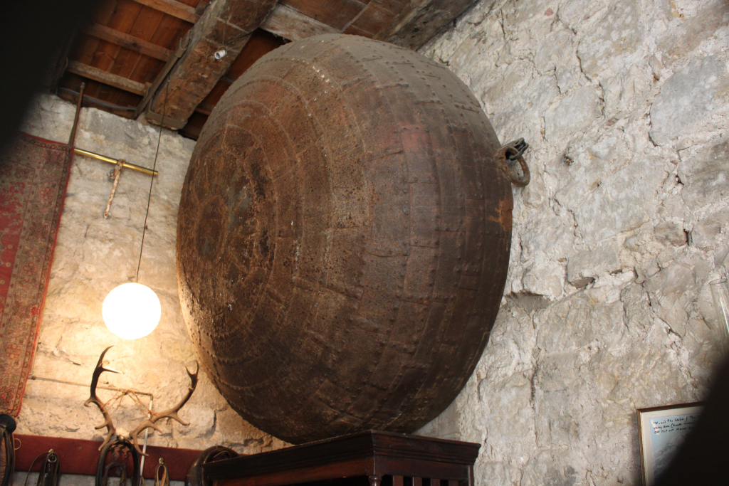 Large, rusty, metal cauldron or pot affixed to a rough stone wall. It appears to be an antique or historical artefact, possibly used for cooking or some other function in the past. The setting seems to be an old building or cellar, with exposed wooden beams and stonework. A simple hanging lamp and a set of antlers on the wall provide context for the age and style of the location.