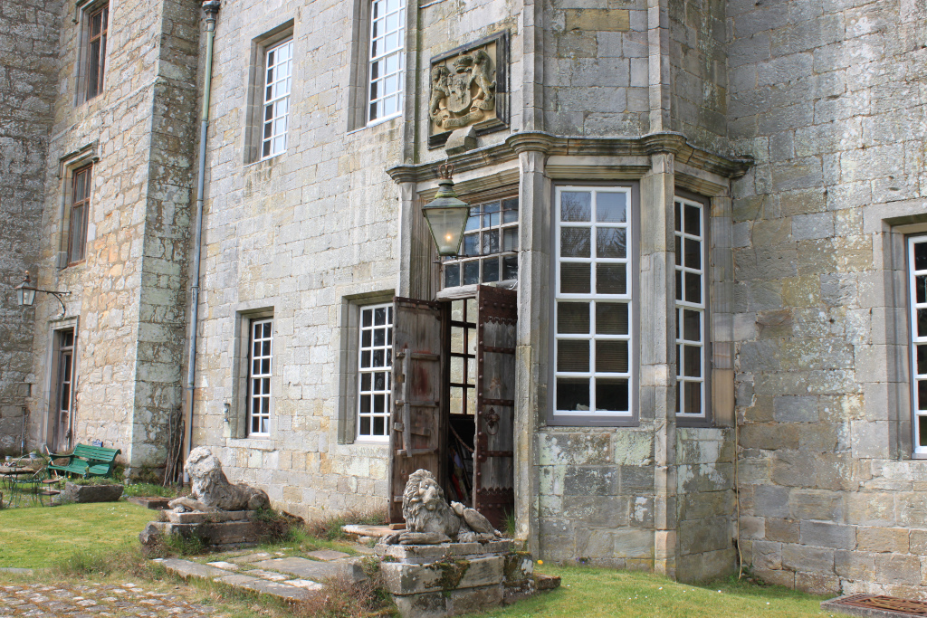 Portion of a stone building, possibly a castle or manor house, with a prominent entrance.  The entrance features a large wooden door, partially open, revealing a glimpse of the interior. Two stone lions flank the entrance.  The building's stonework is aged and shows signs of weathering. The window frames are white and the overall style seems to be historical. A weathered coat of arms is visible above the door. A simple lantern hangs above the entrance. Part of a garden or courtyard is visible in the foreground.