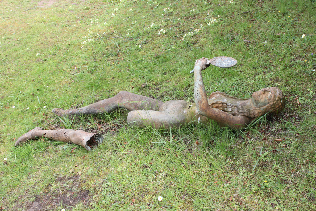 Weathered bronze statue of a nude woman lying on her side in a grassy area. The statue is holding a circular object, possibly a mirror, in one hand. A portion of one leg is broken off and lies separately near the rest of the statue. The overall impression is one of decay and abandonment.