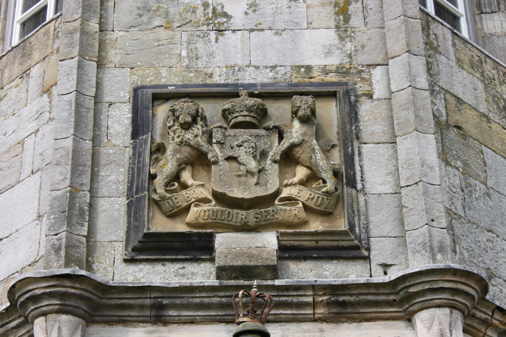 Stone carving of a coat of arms, set into the stonework of a building.  The coat of arms features two lions rampant supporting a shield, with a crown above.  A scroll at the bottom bears an inscription. The stonework shows signs of age and weathering. Below the coat of arms is a smaller, separate stone crown. The overall impression is of an aged, historical building detail.