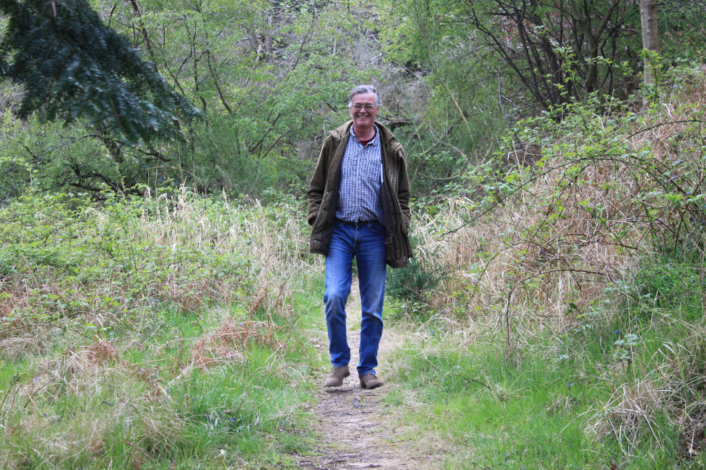 Charlie with short grey hair smiling as he walks down a path in a wooded area. He is wearing a brown jacket, blue jeans, and brown boots. The path is surrounded by green grass and bushes. The overall mood is one of peaceful contentment and enjoyment of nature.