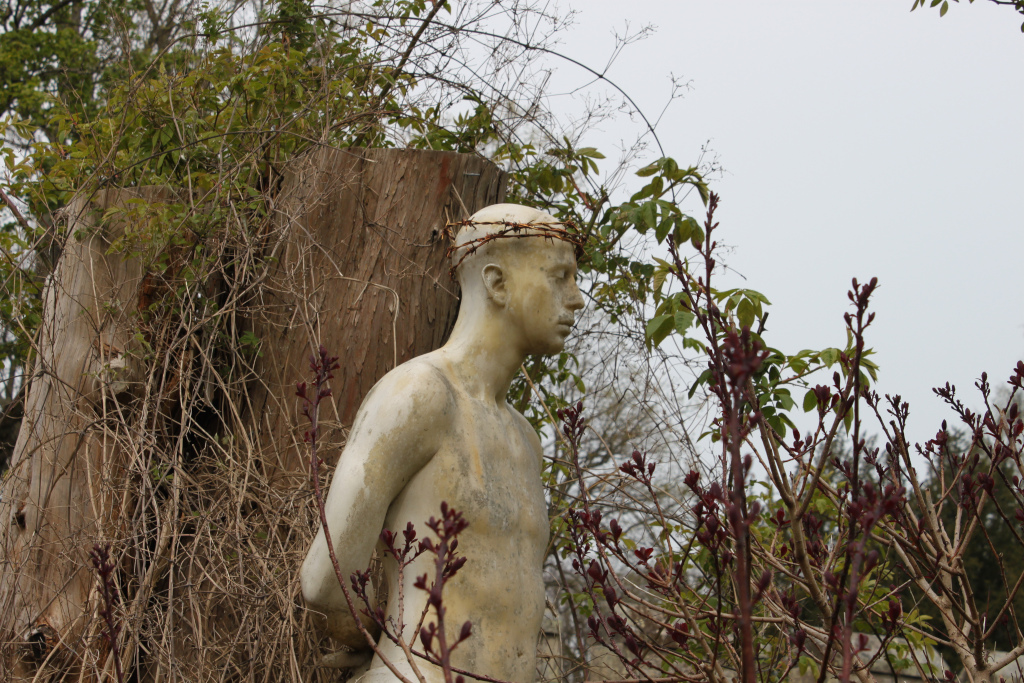 Weathered, light beige statue of a bare-chested man, seemingly made of stone or a similar material. The statue has a crown of barbed wire on its head and is partially obscured by overgrown shrubbery and the remnants of a tree trunk. The overall impression is one of decay, abandonment, and perhaps suffering, suggested by the barbed wire crown.