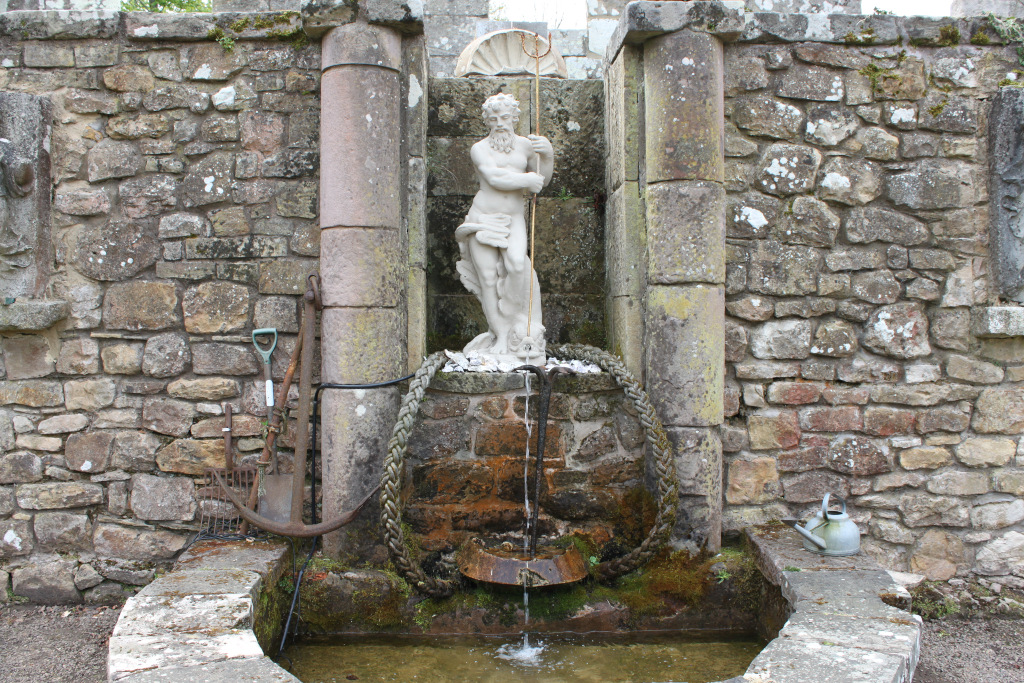 Stone fountain featuring a statue of Neptune, the Roman god of the sea. The statue holds a trident and stands within a niche framed by stone pillars. A small stream of water flows from the base of the statue into a basin below. Around the fountain are various gardening tools and a watering can, suggesting a garden setting. The overall style is classical or perhaps recreating a classical style.
