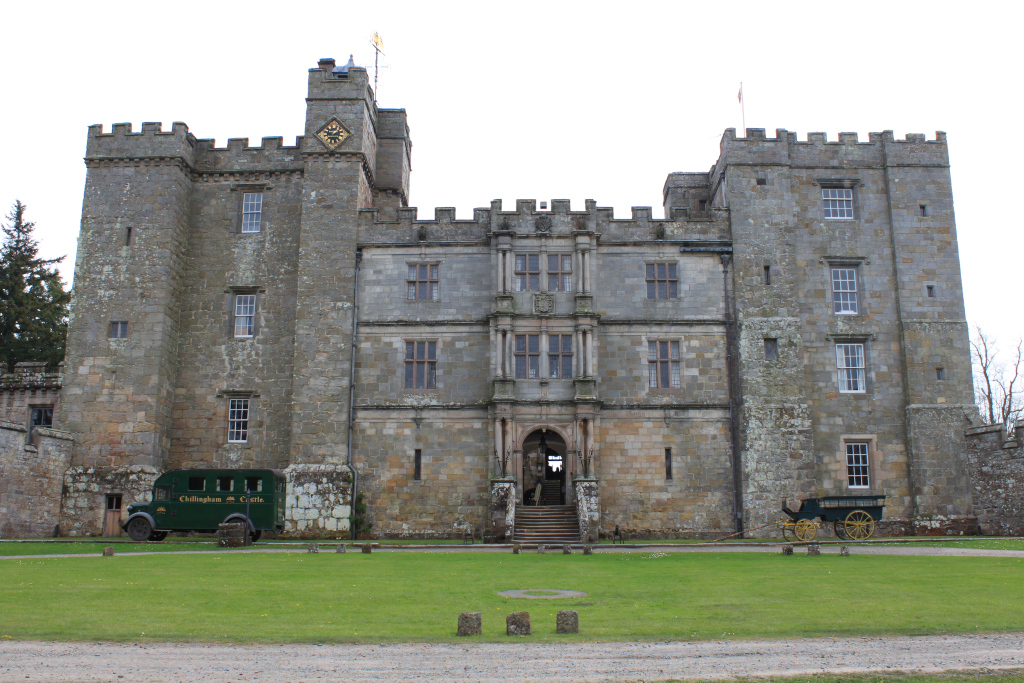 Full frontal view of Chillingham Castle in Northumberland, England. The castle is a large, imposing stone structure with crenellations, multiple towers, and numerous windows.  In the foreground is a neatly kept lawn, and parked near the castle are a vintage green bus and a horse-drawn carriage, suggesting a perhaps tourist-oriented setting.