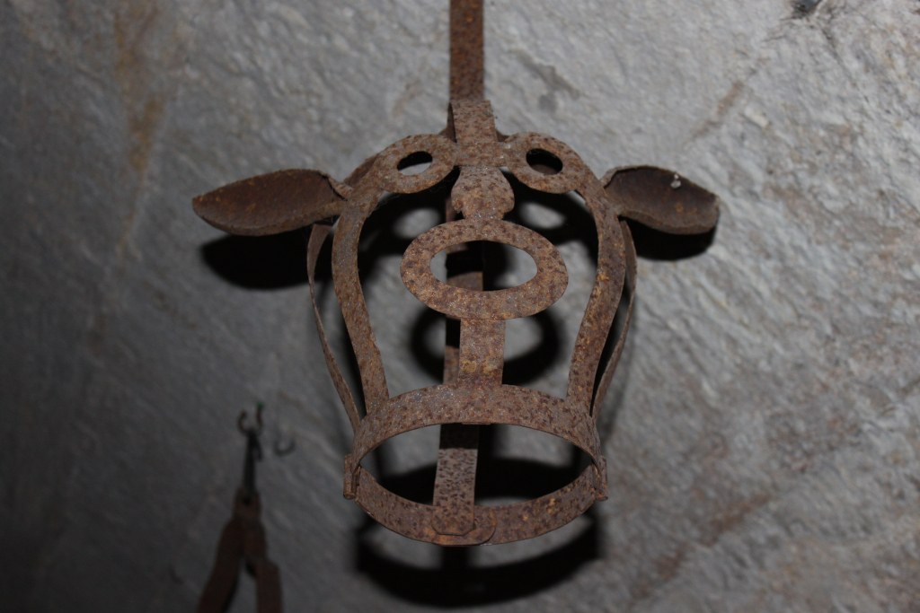 Rusty, metal muzzle, hanging from a ceiling. The muzzle is designed to fit over an animal's head, likely a cow or similar animal, and is heavily corroded with age. The background is a rough, textured stone or concrete wall.