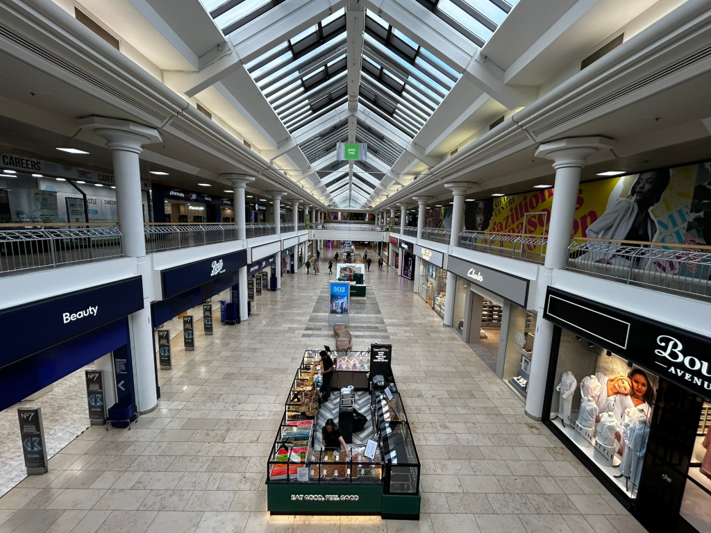 Interior of a largely deserted shopping mall. The high-ceiling space, featuring a glass skylight roof, is spacious but sparsely populated. A few individuals are visible near a small food stall in the centre of the mall. Numerous shops line both sides of the hallway, many displaying advertisements including large posters featuring fashion models. The overall impression is one of quiet emptiness, suggesting a low level of activity or perhaps a period outside of peak shopping hours. The architectural style leans towards modern and clean-lined. The colour palette is muted, with beige stone floors and mostly neutral-toned shopfronts.