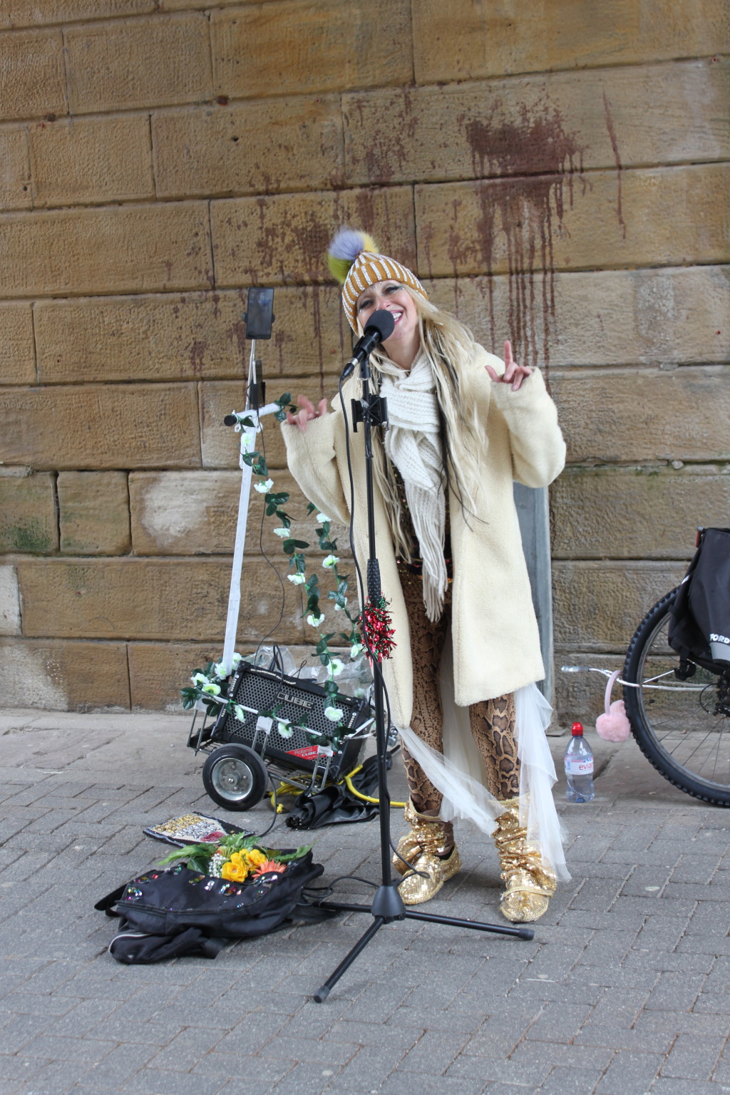 Blonde woman singing into a microphone. She is dressed in a cream-coloured coat, a knitted hat, and patterned leggings. She's wearing glittery gold boots. Her equipment, which includes a small amplifier on a wheeled cart, is decorated with flowers. The background is a stone wall. The overall impression is of a street performer.