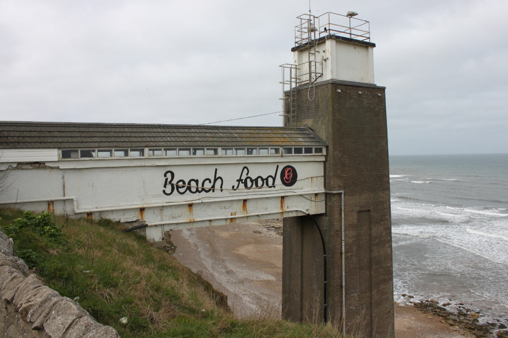Weathered, white structure with the words Beach Food painted on it in black cursive lettering. The structure is built into a cliff overlooking a sandy beach and a choppy sea. A tall, brick tower is attached to the structure, suggesting it may have once served a purpose beyond simply being a food vendor. The overall impression is one of age and coastal erosion.