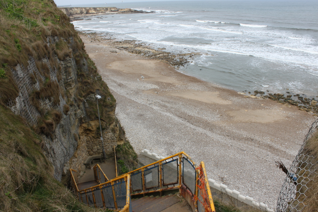 A stairway leading down a cliff face to a beach. The cliff is stabilised with wire mesh, and the stairway has a yellow metal railing. The beach is pebbled, and the ocean is visible in the background with some waves. The overall scene is somewhat desolate and perhaps a little melancholic.