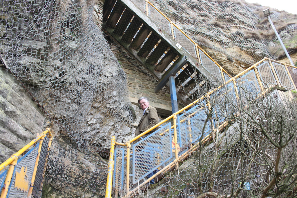 Charlie standing on a steep, outdoor staircase built into a cliff face. The staircase is secured with wire mesh netting to prevent rockfalls, and has yellow and blue metal railings. Charlie is wearing a jacket, and is looking directly at the camera. The overall scene is one of precarious access to what appears to be a structure partially built into the cliff.