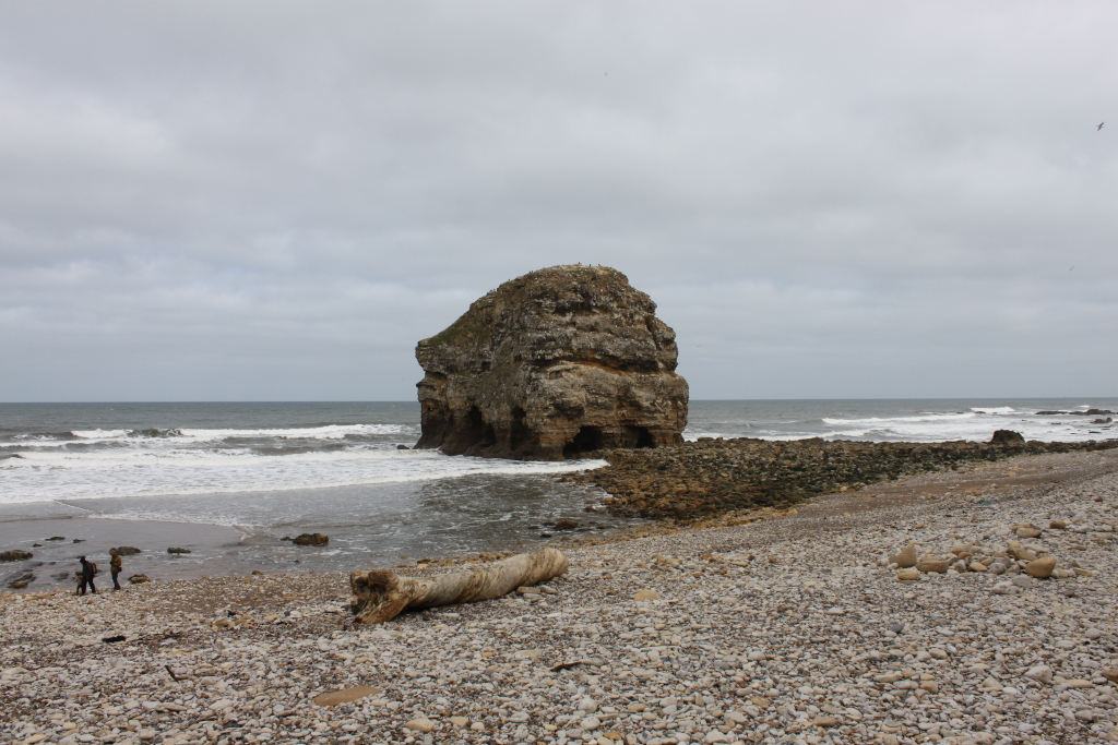 Rocky outcrop in the sea, under a cloudy sky. The outcrop is a significant feature, appearing weathered and eroded with visible caves or hollows. A small pebble beach stretches towards the viewer with a large piece of driftwood lying on the shore. Two figures are visible in the distance on the beach, offering a sense of scale. The overall mood is somewhat bleak and atmospheric due to the grey sky and the solitary nature of the rock formation.
