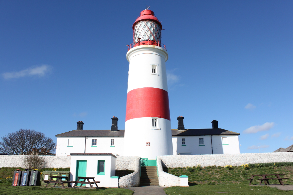Tall, slender lighthouse with a red band around its midsection, standing prominently against a clear blue sky. The lighthouse is attached to a smaller, white keeper's house. The scene is peaceful, with a well-maintained lawn, picnic tables, and recycling bins in the foreground. The overall impression is one of serenity and maritime heritage.