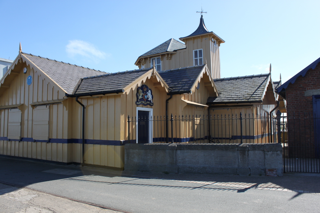 Pale yellow building with a dark grey slate roof, situated on a sunny day. The building appears to be made of wood and features decorative wooden trim. It has a taller, central section that resembles a small tower. A low wall and black metal fence partially enclose the building. The central section of the building displays a crest or coat of arms. The style suggests a historical or possibly nautical context, perhaps a boathouse or similar structure.