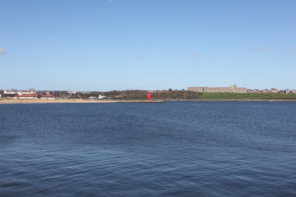 Wide shot of a calm sea. In the background, a sandy beach stretches along the shoreline, with buildings and a residential area visible beyond. A striking red lighthouse is situated on a pier, prominently featured in the middle ground. The sky is a clear, bright blue, indicating a sunny day. The overall impression is one of tranquillity and serenity, capturing a coastal scene on a clear day.