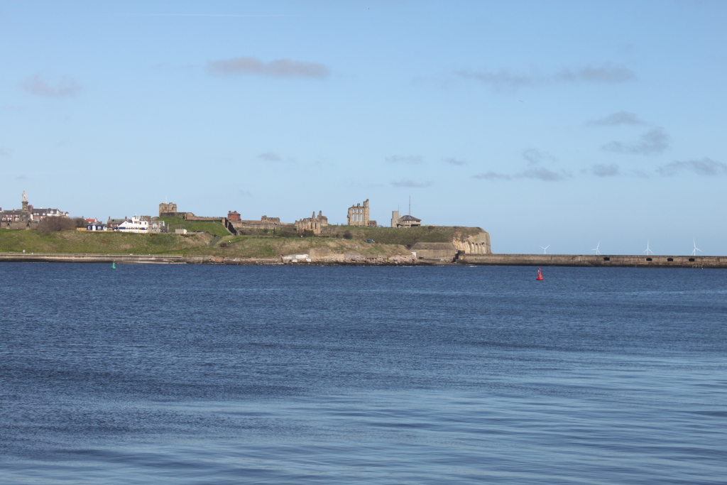 Long shot of a coastline on a sunny day. In the background, there is a line of buildings and structures, including what appears to be the ruins of a priory, sitting atop a cliff. To the right, there are several wind turbines visible in the distance. The foreground is dominated by calm, blue water. The overall atmosphere is peaceful and serene.