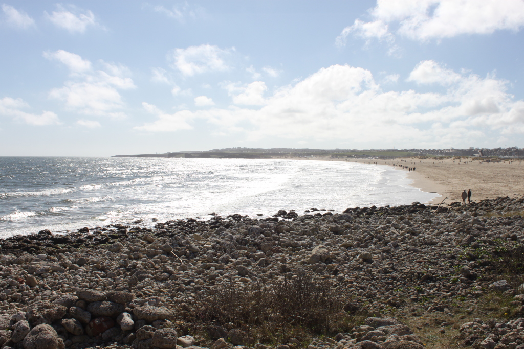 Long, sandy beach stretching from the rocky foreground to the horizon, where a line of buildings or houses is visible. The beach is sparsely populated with people visible in the distance. The ocean is relatively calm with small waves breaking on the shore. The foreground is dominated by a rocky, uneven terrain composed of various sized grey stones. The sky is mostly clear, with scattered, fluffy clouds.