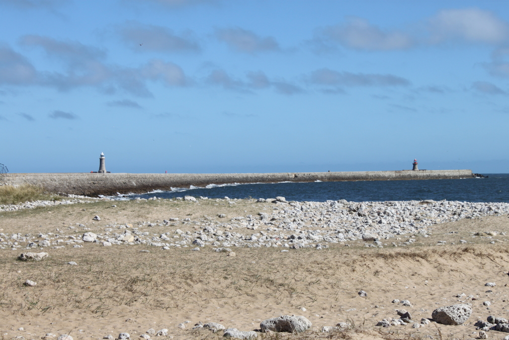 Long breakwater stretching across the ocean under a mostly clear blue sky. Two lighthouses are visible, one at either end of the breakwater. The foreground consists of a sandy beach strewn with light-coloured rocks. The overall impression is one of a tranquil coastal scene.