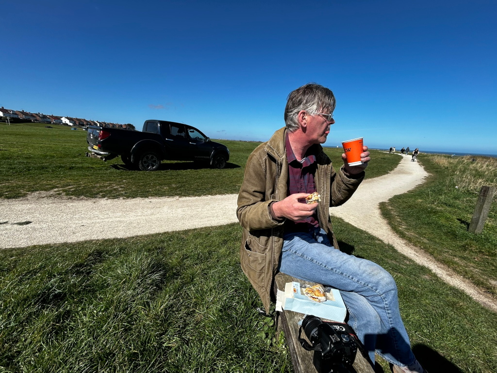 Charlie sitting on a bench outdoors, enjoying a snack and a drink. He's wearing a brown jacket and jeans. In the background, a blue pickup truck is parked on a grassy area, and a path leads towards clear blue sky. The overall scene suggests a relaxed coastal, setting on a sunny day
